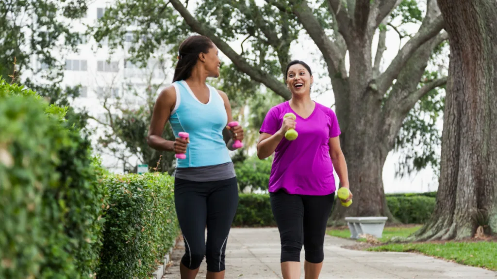 Two women walking outdoors on a tree-lined path, holding small hand weights and smiling as they engage in conversation while getting their 10,000 steps a day.